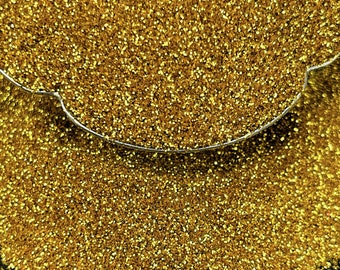 May include: Close-up of a pile of gold glitter. The glitter is densely packed, reflecting light and creating a sparkling effect. A thin, curved, silver wire is partially visible at the top of the image.
