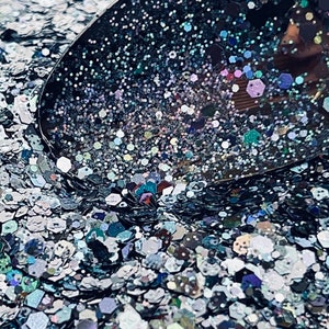 May include: Close-up of a pile of iridescent glitter in various shapes and sizes. The glitter is primarily silver, with hints of blue, green, and purple. A dark, curved object is partially visible, reflecting the glitter.