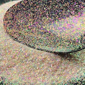 Puede incluir: Primer plano de purpurina iridiscente en una cuchara y esparcida sobre una superficie. La purpurina tiene una mezcla de colores, incluyendo rosa, morado y verde, creando un efecto brillante. La purpurina es fina y parece ser de grado cosmético.