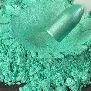 May include: Close-up of a pile of shimmering, teal-colored powder. A nail polish swatch displays the same color, showcasing its metallic finish. The powder appears finely milled, suggesting a cosmetic product.