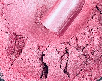 May include: Close-up of a pink metallic nail polish sample on a white stick, set against a backdrop of loose, shimmering pink powder. The nail polish has a smooth, reflective surface, and the powder has a textured appearance.