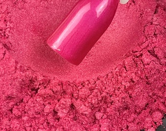 May include: A close-up of a vibrant pink nail polish swatch on a white stick, set against a backdrop of loose, finely milled pink powder. The nail polish has a glossy, reflective finish, showcasing a rich, saturated color.