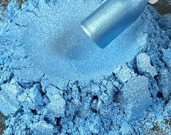 Puede incluir: Primer plano de un montón de polvo azul claro con brillo metálico. Una muestra con un acabado metálico azul a juego se coloca en el polvo. El polvo tiene un aspecto texturizado y granulado, lo que sugiere un producto cosmético.