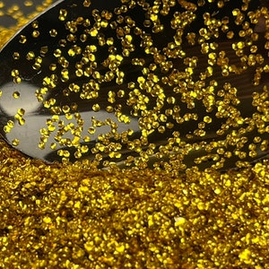 Könnte beinhalten: Nahaufnahme eines glänzenden, silbernen Löffels, gefüllt mit goldenem Glitzer. Der Glitzer ist auf einer dunklen Oberfläche verstreut und erzeugt einen funkelnden Effekt. Der Glitzer besteht aus kleinen, unregelmäßigen Formen.