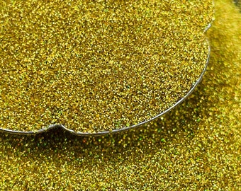 May include: Close-up of a pile of gold glitter, with a silver-colored scoop partially visible. The glitter is finely milled and reflects light, creating a sparkling effect. The background is dark, making the glitter stand out.