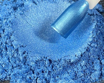 Puede incluir: Primer plano de un montón de polvo azul vibrante con brillo metálico. Una muestra de uña de plástico blanco muestra un esmalte de uñas azul a juego, mostrando el color y el acabado del producto. El polvo parece finamente molido, lo que sugiere un cosmético o un suministro de arte.