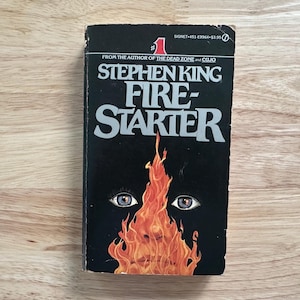 May include: A mass market paperback book titled Firestarter by Stephen King with a black cover featuring a graphic illustration of a face partially obscured by orange and yellow flames and two visible eyes centered above the fire.