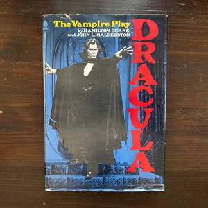 May include: A book titled "The Vampire Play" by Hamilton Deane and John L. Balderston. The cover features a man in a black cape and a red title "Dracula" on the right side.