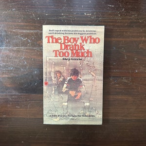 May include: A paperback book cover with a red, white, and blue color scheme. The title is "The Boy Who Drank Too Much" by Shep Greene. The cover features a drawing of a young boy sitting on a bench with a bottle in his hand.