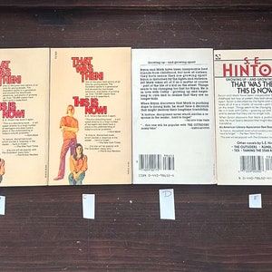 May include: A collection of six paperback books, all with similar covers featuring a yellow background with red and black text. The books are titled "That Was Then, This Is Now" and "The Outsiders" by S.E. Hinton. The books are arranged in a row on a wooden surface.