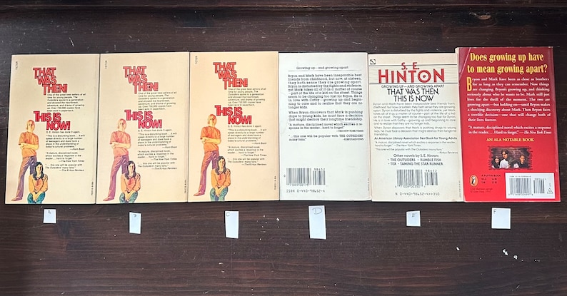May include: A collection of six paperback books, all with similar covers featuring a yellow background with red and black text. The books are titled "That Was Then, This Is Now" and "The Outsiders" by S.E. Hinton. The books are arranged in a row on a wooden surface.
