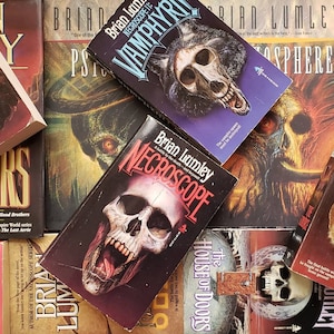 May include: A collection of paperback books with horror themes. The books are stacked and arranged in a pile. The covers feature various illustrations of monsters, skulls, and other spooky imagery. The titles of the books include "Deadspawn", "Bloodwars", "Vamphyr", "Necroscope", "The Source", and "The House of Doors".