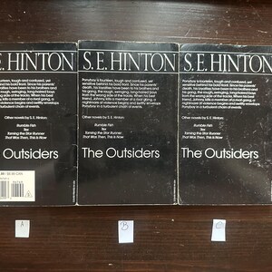 May include: Three paperback copies of the book "The Outsiders" by S.E. Hinton. The books are black with white text and have a price tag on the bottom left corner.