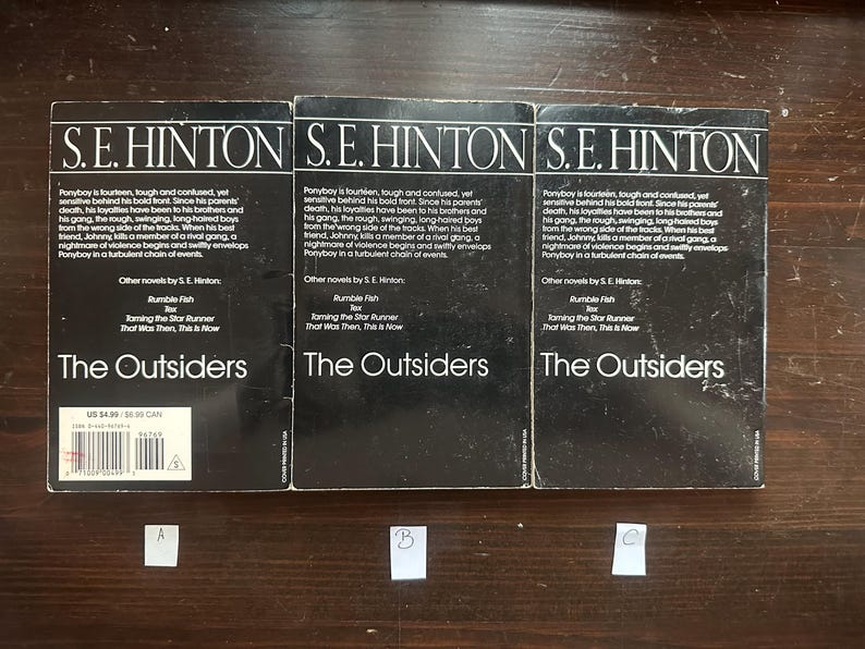May include: Three paperback copies of the book "The Outsiders" by S.E. Hinton. The books are black with white text and have a price tag on the bottom left corner.