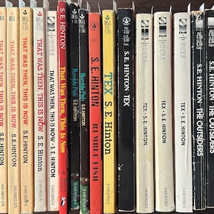 May include: A shelf of paperback books by S.E. Hinton, including titles like "That Was Then, This Is Now", "Rumble Fish", and "Tex".