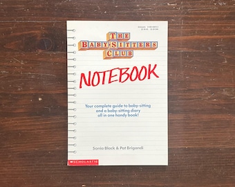 May include: A spiral-bound notebook with the title "THE BABYSITTERS CLUB NOTEBOOK" in red and yellow lettering. The notebook includes the text "Your complete guide to baby-sitting and a baby-sitting diary all in one handy book!" on a wooden background.