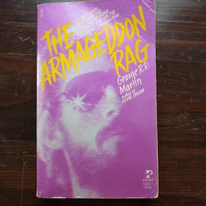 May include: A paperback book cover with a purple background and yellow text. The title is "The Armageddon Rag" by George R.R. Martin. The book is a pocket edition and costs $3.95.
