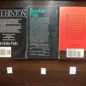 May include: Three paperback books with red, black, and white covers. The books are titled "Rumble Fish" by S.E. Hinton. The back covers feature text about the books and the author.