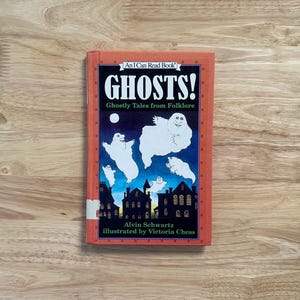 May include: A hardcover book titled Ghosts! Ghostly Tales from Folklore by Alvin Schwartz, illustrated by Victoria Chess. The orange cover features a dark blue night scene with white cartoon ghosts floating above a silhouette of dark buildings.