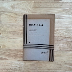 May include: A tan paperback play script titled Dracula, freely adapted by Crane Johnson from the novel by Bram Stoker. The cover features black vertical and horizontal stripes, two small stars, and the text Dramatists Play Service Inc.