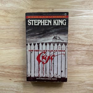 May include: A mass market paperback book titled Cujo by Stephen King. The cover features a white picket fence with red blood-like drips, a dark stormy sky, and a house in the distance. Text includes: See the blood-chilling new film from Warner Bros.