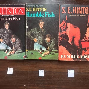 May include: Three paperback copies of the book Rumble Fish by S.E. Hinton. The book covers feature a red, white, and blue color scheme with a pool table and people playing pool.