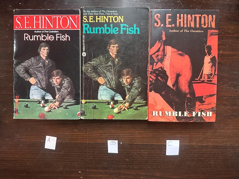 May include: Three paperback copies of the book Rumble Fish by S.E. Hinton. The book covers feature a red, white, and blue color scheme with a pool table and people playing pool.
