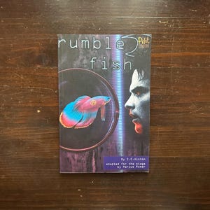 May include: A book cover with a purple background and a fish in a bowl. The title of the book is "rumble fish" and the author is S-C-Hinton. The book was adapted for the stage by Marcus Romer.