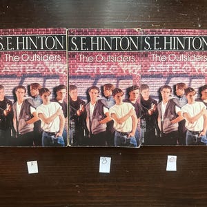 May include: Three paperback copies of the book "The Outsiders" by S.E. Hinton. The books are arranged side-by-side on a wooden surface. Each book cover features a group of teenagers standing in front of a brick wall with graffiti.