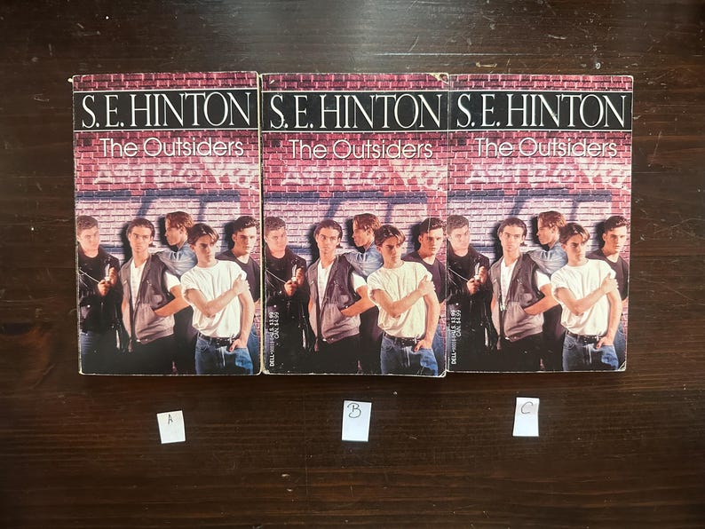 May include: Three paperback copies of the book "The Outsiders" by S.E. Hinton. The books are arranged side-by-side on a wooden surface. Each book cover features a group of teenagers standing in front of a brick wall with graffiti.