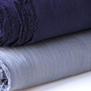 May include: Close-up of two rolled textiles. The top textile is a deep indigo blue with a fringed edge. The bottom textile is a light blue-grey color. Both textiles appear to be made of a woven fabric.