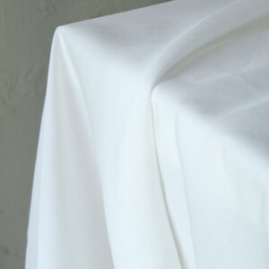 Pode incluir: Um close-up de tecido branco drapeado sobre uma superf&iacute;cie. O tecido &eacute; liso e parece ser um material leve, com dobras e sombras suaves. O fundo &eacute; cinza neutro.