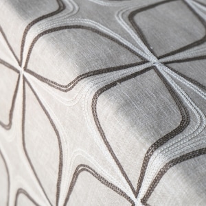 Può includere: Primo piano di un tessuto grigio chiaro con un motivo geometrico ripetuto. Il motivo è creato con cuciture grigio scuro e bianche, che formano linee e forme curve. Il tessuto ha un aspetto strutturato.