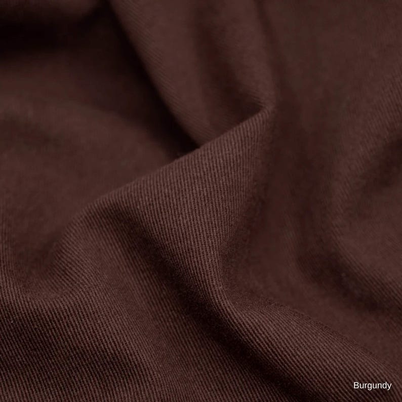 K&ouml;nnte beinhalten: Ein dunkelbrauner Stoff mit einem sichtbaren diagonalen K&ouml;perbindungsmuster. Das Material ist gefaltet, wodurch weiche Schatten und Lichter auf der Oberfl&auml;che entstehen. Das Wort Burgundy ist in der unteren rechten Ecke abgedruckt.