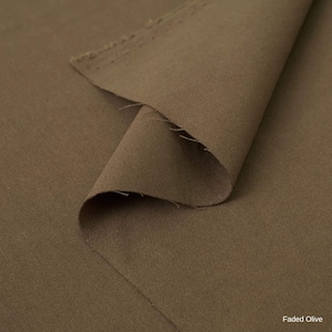 K&ouml;nnte beinhalten: Ein St&uuml;ck Stoff in einem ged&auml;mpften, erdigen Farbton von verblasstem Olivgr&uuml;n. Das Material weist eine glatte, matte Textur mit einem sichtbaren Webmuster auf. Eine Ecke des Stoffes ist umgeschlagen und zeigt die rohe, leicht ausgefranste Kante.