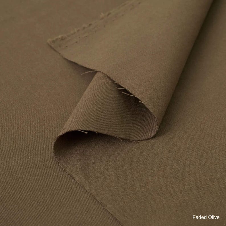 K&ouml;nnte beinhalten: Ein St&uuml;ck Stoff in einem ged&auml;mpften, erdigen Farbton von verblasstem Olivgr&uuml;n. Das Material weist eine glatte, matte Textur mit einem sichtbaren Webmuster auf. Eine Ecke des Stoffes ist umgeschlagen und zeigt die rohe, leicht ausgefranste Kante.
