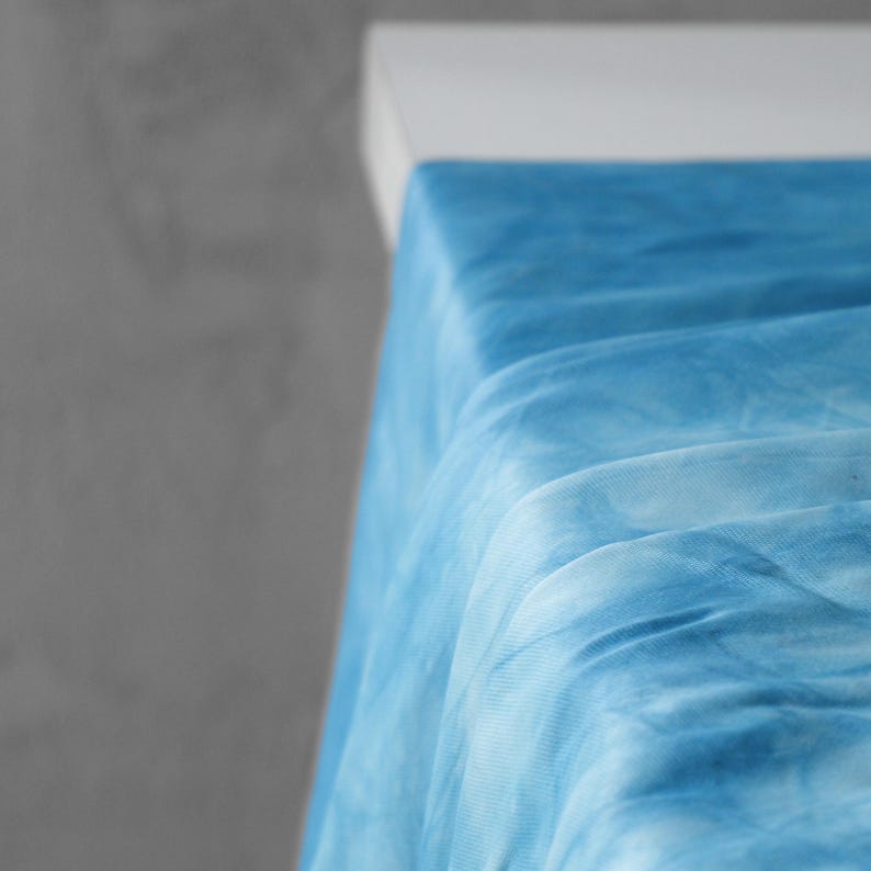 Puede incluir: Primer plano de una tela azul te&ntilde;ida con efecto tie-dye, drapeada sobre una superficie blanca. La tela tiene una textura suave y fluida con diferentes tonos de azul y blanco, creando un efecto acuarela. La tela se utiliza probablemente para ropa o decoraci&oacute;n del hogar.