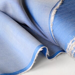 May include: Close-up of a light blue fabric with a subtle texture. The fabric is folded, revealing layers of varying shades of blue. The edges are frayed, showing the raw texture of the material. A darker blue border trims the fabric.