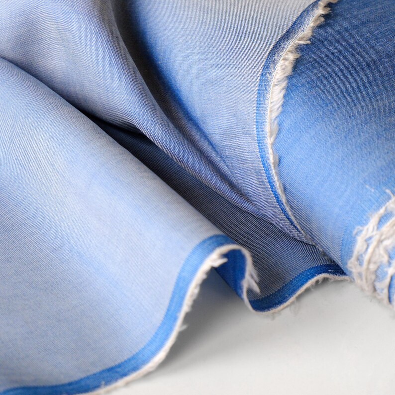 May include: Close-up of a light blue fabric with a subtle texture. The fabric is folded, revealing layers of varying shades of blue. The edges are frayed, showing the raw texture of the material. A darker blue border trims the fabric.