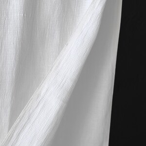 May include: Close-up of white fabric with a textured, slightly wrinkled appearance. The fabric is folded, creating diagonal lines and shadows against a black background. The material appears to be lightweight and possibly linen or cotton.