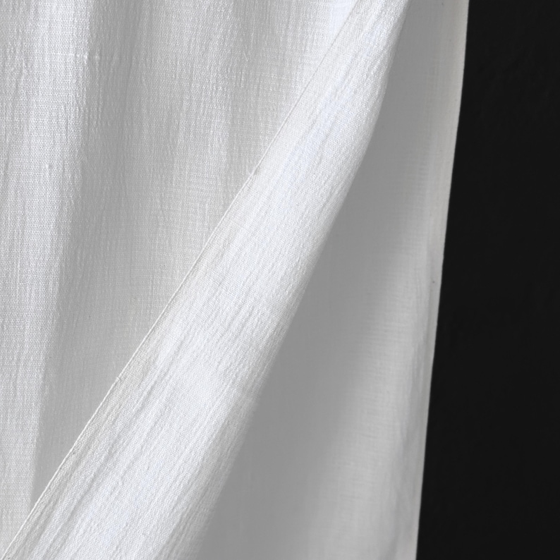 May include: Close-up of white fabric with a textured, slightly wrinkled appearance. The fabric is folded, creating diagonal lines and shadows against a black background. The material appears to be lightweight and possibly linen or cotton.