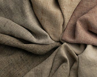May include: A collection of folded fabric swatches featuring a variety of neutral earth tones including beige, tan, taupe, brown, and muted rose. The material displays a visible woven texture with a coarse, natural grain throughout the surface.