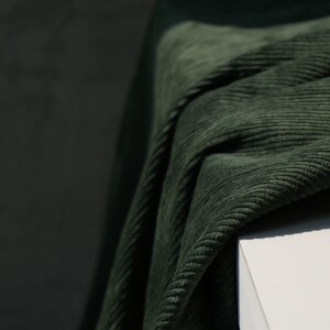 May include: Close-up of a dark green corduroy fabric draped over a white surface. The fabric has a ribbed texture and is folded, creating shadows and highlights. The color is a deep, muted green.