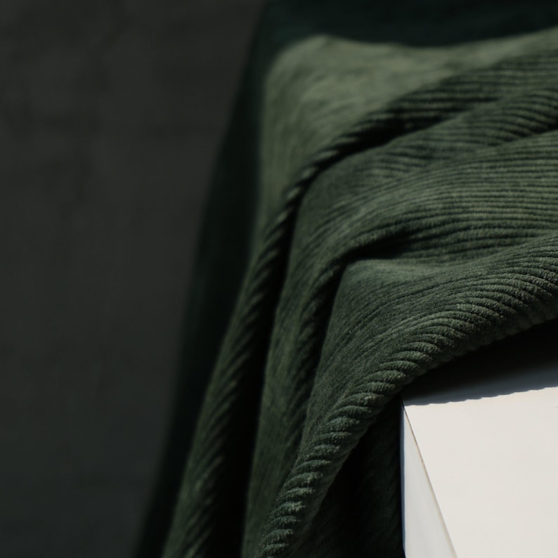 May include: Close-up of a dark green corduroy fabric draped over a white surface. The fabric has a ribbed texture and is folded, creating shadows and highlights. The color is a deep, muted green.