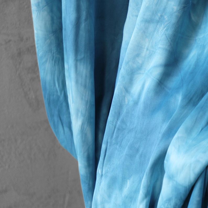 Puede incluir: Primer plano de una tela fluida con un estampado tie-dye en tonos azules. La tela est&aacute; drapeada, creando pliegues y sombras suaves. El patr&oacute;n de tinte tiene un efecto acuarela, con &aacute;reas m&aacute;s claras y oscuras.