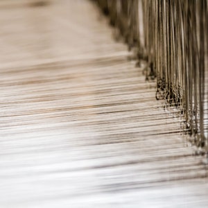 May include: Close-up of a loom with many threads running parallel to each other. The threads are white and the loom is made of metal.