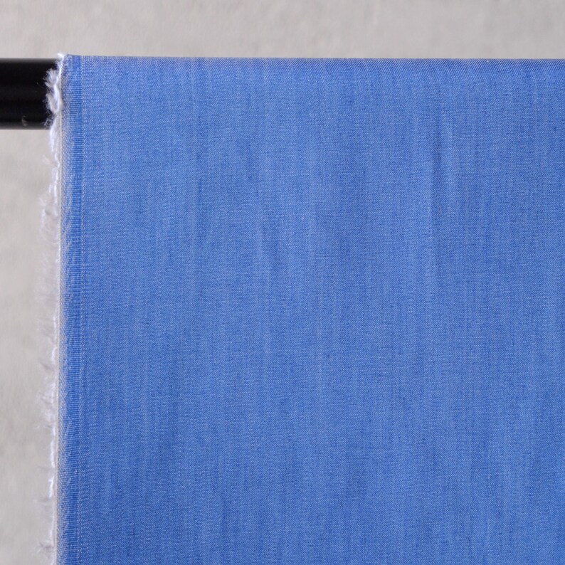 May include: A close-up of a section of blue fabric. The fabric is a solid, light blue colour with a slightly textured appearance. The edge of the fabric is frayed, revealing the raw edge of the material. The fabric is draped over a black rod.