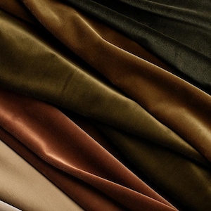 May include: A collection of folded velvet fabric in various earth tones, including shades of beige, rust orange, olive green, and dark forest green. The material features a soft, dense pile with visible light reflections and shadows across the folds.