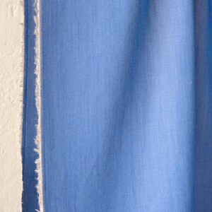 May include: Close-up of a light blue fabric with a textured appearance. The fabric is next to a white wall with a frayed edge. The fabric is a solid colour and appears to be a textile.