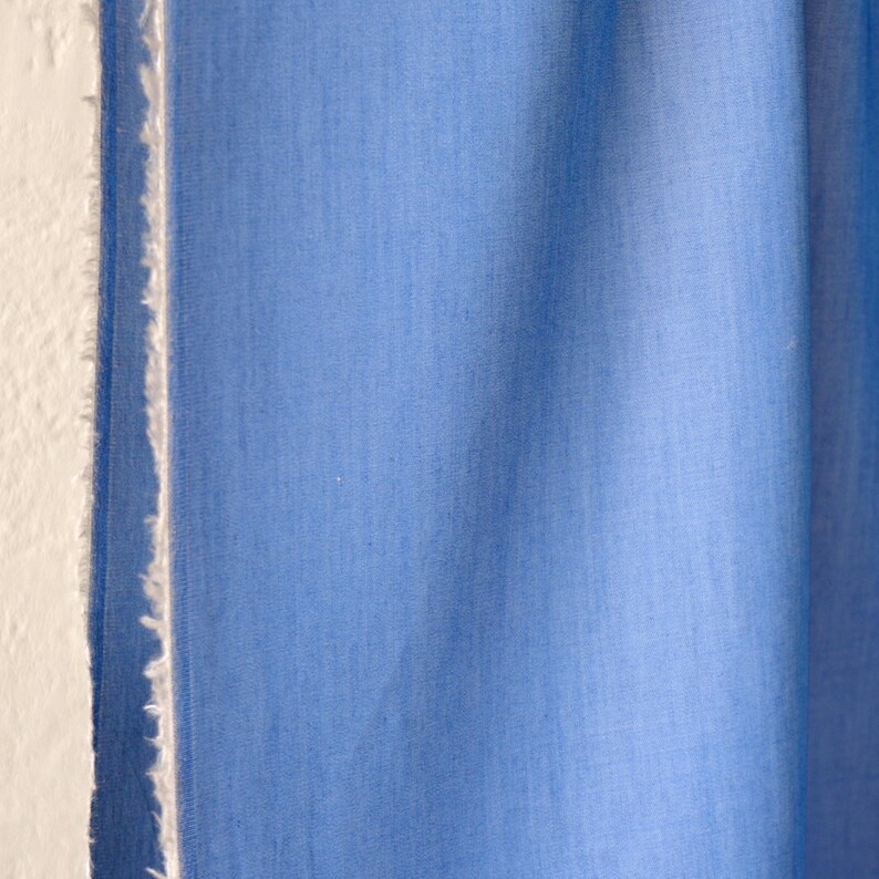 May include: Close-up of a light blue fabric with a textured appearance. The fabric is next to a white wall with a frayed edge. The fabric is a solid colour and appears to be a textile.